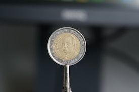 A close-up image of a coin held by a clamp-like tool. The coin appears to be a Spanish Euro, featuring a portrait in the center and surrounded by stars. The background is blurred, emphasizing the focus on the coin.