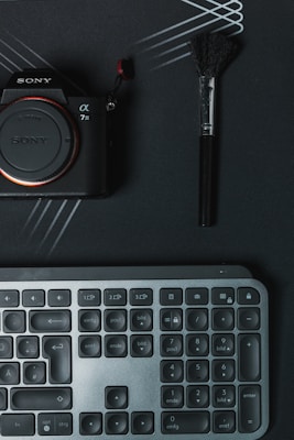 A digital camera with the brand name partially visible, a makeup brush, and a keyboard are arranged on a dark surface with intersecting lines. The camera is on the left, the brush on the right, and the keyboard below.