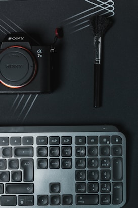 A digital camera with the brand name partially visible, a makeup brush, and a keyboard are arranged on a dark surface with intersecting lines. The camera is on the left, the brush on the right, and the keyboard below.