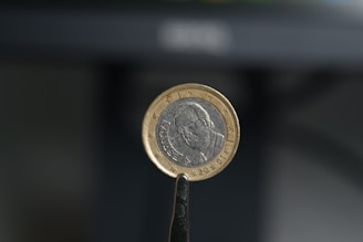 A close-up view of a euro coin held between metal tweezers. The coin displays a portrait on one side with the word 'España' inscribed. The background is blurred, emphasizing the focus on the coin.