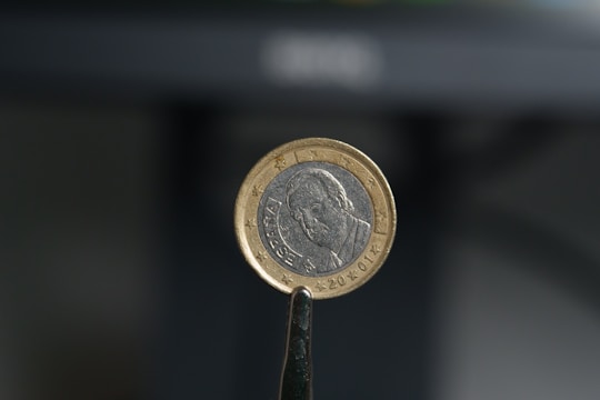A close-up view of a euro coin held between metal tweezers. The coin displays a portrait on one side with the word 'España' inscribed. The background is blurred, emphasizing the focus on the coin.