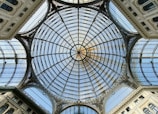 An intricate glass dome ceiling with elaborate metal framework, forming a geometric pattern. The structure features a central circular design surrounded by radiating lines and arches, framed by ornate architectural elements.
