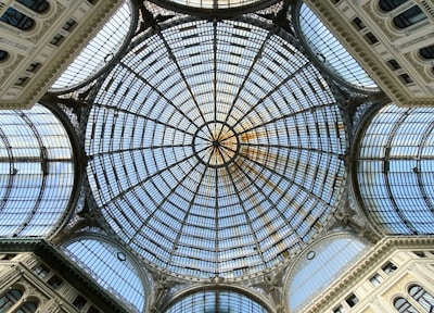An intricate glass dome ceiling with elaborate metal framework, forming a geometric pattern. The structure features a central circular design surrounded by radiating lines and arches, framed by ornate architectural elements.