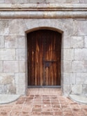 A tall wooden door with a rustic appearance is set into a stone wall. The door features vertical wooden planks with visible grain patterns and is secured by a simple metal lock. The surrounding wall is constructed from large, weathered stone blocks, adding to the aged and sturdy appearance. The ground in front is paved with red brick tiles.