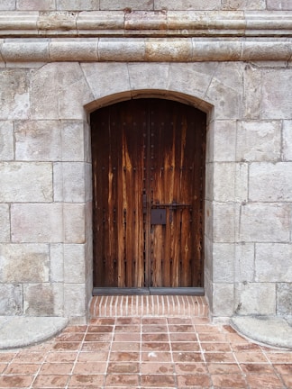 A tall wooden door with a rustic appearance is set into a stone wall. The door features vertical wooden planks with visible grain patterns and is secured by a simple metal lock. The surrounding wall is constructed from large, weathered stone blocks, adding to the aged and sturdy appearance. The ground in front is paved with red brick tiles.