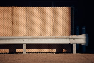 A metal guardrail stands in front of a tan-colored chain-link fence. The scene is sparsely lit, casting strong shadows on the ground. The textures of the metal and the fence provide a contrast, with the guardrail appearing rusted and weathered.
