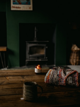 A cozy scene with a wooden table in the foreground, featuring a knit sweater draped over it. A lit candle in a small glass jar adds a warm glow to the setting. A ceramic mug is placed closer to the edge of the table. In the background, a wood-burning stove is visible, creating an inviting atmosphere.