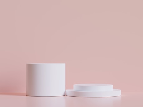 Two smooth, cylindrical, white podiums or platforms are arranged on a soft pink surface with a matching pink background. The scene is minimalistic and clean, focusing on the geometric simplicity of the objects.