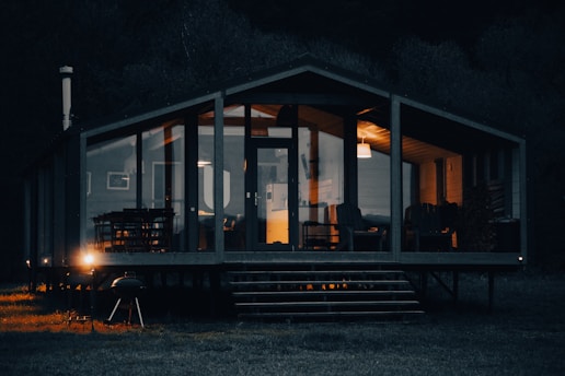 A cozy cabin with large glass windows illuminated warmly from the inside, set against a dark, wooded background. The cabin's interior shows comfortable chairs and a dining table, while the exterior features a barbecue grill near the porch.
