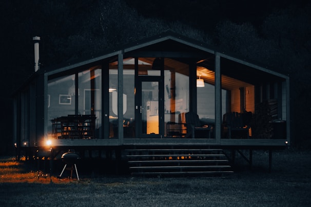 A cozy cabin with large glass windows illuminated warmly from the inside, set against a dark, wooded background. The cabin's interior shows comfortable chairs and a dining table, while the exterior features a barbecue grill near the porch.