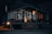 A cozy cabin with large glass windows illuminated warmly from the inside, set against a dark, wooded background. The cabin's interior shows comfortable chairs and a dining table, while the exterior features a barbecue grill near the porch.