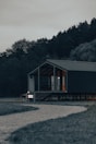 A modern cabin with a number 101 displayed outside stands in a serene forest setting. The cabin has large glass windows, through which warm lights from inside are visible. A winding gravel path leads up to the porch, surrounded by lush green grass and dense trees.