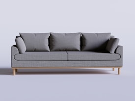 A modern, minimalist grey sofa with several large cushions on a light wooden floor. The overall design is sleek and simple, with clean lines that contribute to a contemporary look.