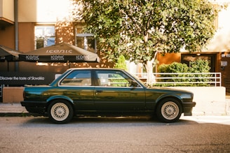 A classic dark green car is parked on a street with a tree nearby, casting shadows on the ground. Behind the car, a building with large windows is visible, featuring outdoor umbrellas with the word 'ICONIC' printed on them. There is greenery beside the building, and the sunlight creates a warm atmosphere.