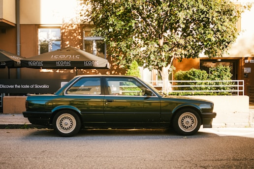 A classic dark green car is parked on a street with a tree nearby, casting shadows on the ground. Behind the car, a building with large windows is visible, featuring outdoor umbrellas with the word 'ICONIC' printed on them. There is greenery beside the building, and the sunlight creates a warm atmosphere.