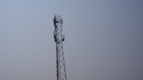 A tall metal telecommunications tower stands against a clear blue sky. The tower features multiple antennas attached along its sides, indicating its use for communication purposes.