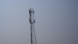 A tall metal telecommunications tower stands against a clear blue sky. The tower features multiple antennas attached along its sides, indicating its use for communication purposes.