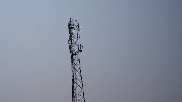 A tall metal telecommunications tower stands against a clear blue sky. The tower features multiple antennas attached along its sides, indicating its use for communication purposes.