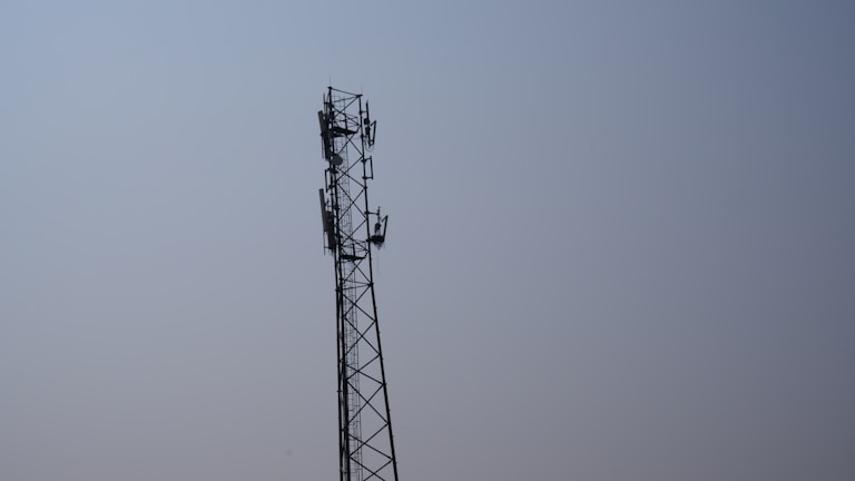 A tall metal telecommunications tower stands against a clear blue sky. The tower features multiple antennas attached along its sides, indicating its use for communication purposes.