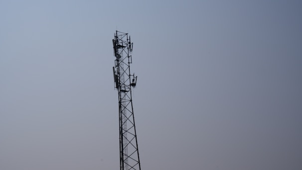 A tall metal telecommunications tower stands against a clear blue sky. The tower features multiple antennas attached along its sides, indicating its use for communication purposes.