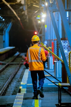 A worker in an orange safety vest and hard hat walks along a platform inside an industrial or rail setting. The area is dimly lit with artificial lights. Tracks run parallel to the platform, and in the background, other workers can be seen attending to their tasks.