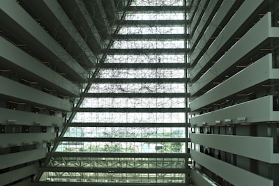 The image captures the interior of a large, modern building with multiple floors visible. The walls on either side are lined with balconies and corridors, suggesting a hotel or commercial space. The central area features a complex network of metal or wire structures, creating an intricate pattern. Natural light floods in, illuminating the spacious atrium.