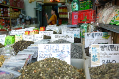 Various types of tea leaves are displayed in containers with labels written in a foreign language, likely Thai. The tea is accompanied by other packaged goods lining the shelves in a small, busy shop with colorful packaging. A person is seen in the background holding a smartphone.