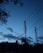 Telecommunication towers stand silhouetted against a darkening sky with scattered clouds. Surrounding trees and foliage are visible, creating a serene, evening landscape. Power lines stretch diagonally across the scene, contributing to the industrial feel.
