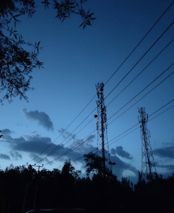 Telecommunication towers stand silhouetted against a darkening sky with scattered clouds. Surrounding trees and foliage are visible, creating a serene, evening landscape. Power lines stretch diagonally across the scene, contributing to the industrial feel.