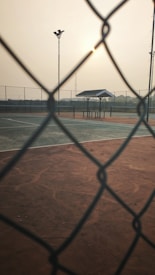 A tennis court is viewed through a chain-link fence, with a pavilion in the background. The setting sun casts a warm glow over the scene, and there are tall floodlights visible under a hazy sky.