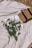 A beige blanket is spread on the ground with a wedding magazine featuring a bride on the cover placed on it. Beside the magazine is a woven straw bag with black straps. A bouquet of white flowers and a bottle of what appears to be a skincare or cosmetic product are also positioned on the blanket.