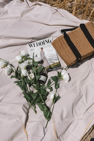 A beige blanket is spread on the ground with a wedding magazine featuring a bride on the cover placed on it. Beside the magazine is a woven straw bag with black straps. A bouquet of white flowers and a bottle of what appears to be a skincare or cosmetic product are also positioned on the blanket.