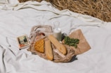 A picnic setup on a white blanket with various items including a book, a lattice-top pie, baguettes wrapped in brown paper, a bunch of green grapes on a paper wrapper, and a glass bottle filled with milk. There is dried grass partially visible in the background.