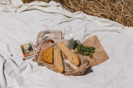 A picnic setup on a white blanket with various items including a book, a lattice-top pie, baguettes wrapped in brown paper, a bunch of green grapes on a paper wrapper, and a glass bottle filled with milk. There is dried grass partially visible in the background.