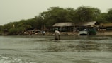 A riverside scene featuring several people engaged in various activities near the shore. There are boats docked at the water's edge, and a large makeshift structure with a thatched roof is visible. The area is surrounded by lush green trees, and a national flag is prominently displayed.