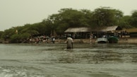 A riverside scene featuring several people engaged in various activities near the shore. There are boats docked at the water's edge, and a large makeshift structure with a thatched roof is visible. The area is surrounded by lush green trees, and a national flag is prominently displayed.