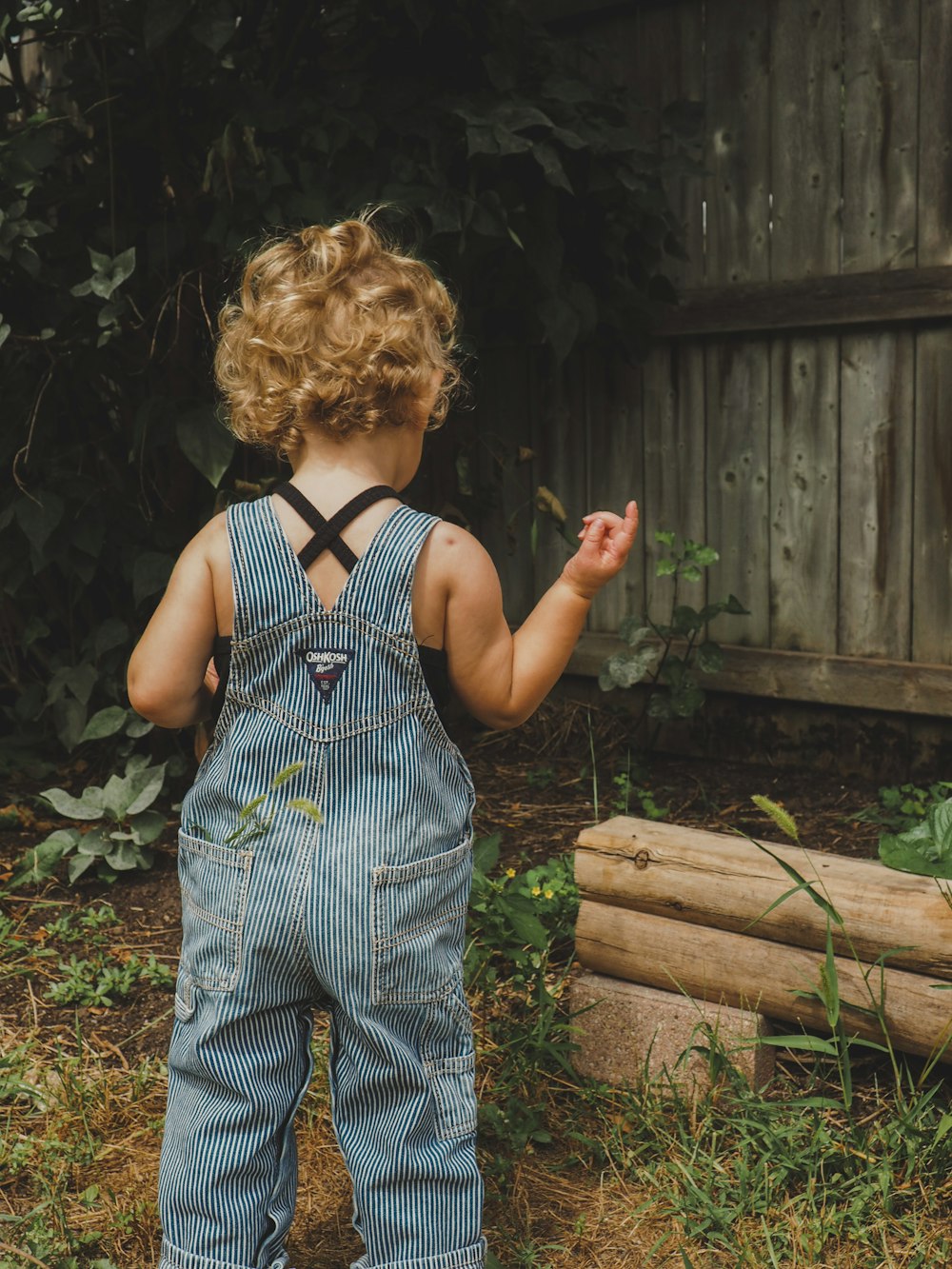 a toddler in overalls standing in front of a fence
