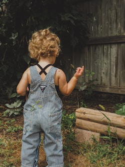 A young child with curly blond hair stands outdoors, facing away, wearing striped denim overalls. The child is surrounded by greenery and a wooden fence, suggesting a garden or backyard setting.