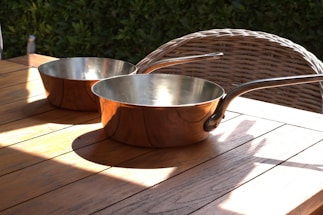 Two copper pans with long handles are placed on a wooden table. The table is outdoors, situated next to a wicker chair with a backdrop of green foliage. Sunlight casts shadows and highlights over the wood and metal surfaces.