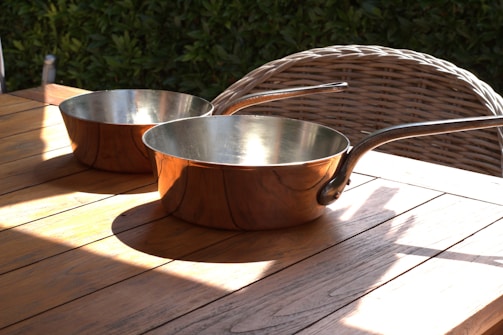 Two copper pans with long handles are placed on a wooden table. The table is outdoors, situated next to a wicker chair with a backdrop of green foliage. Sunlight casts shadows and highlights over the wood and metal surfaces.