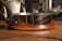 A copper pot with a dark metal handle sits on a wooden surface, reflecting sunlight. The handle is secured with metal rivets, and in the background, there are blurred potted plants with green leaves and pink flowers.