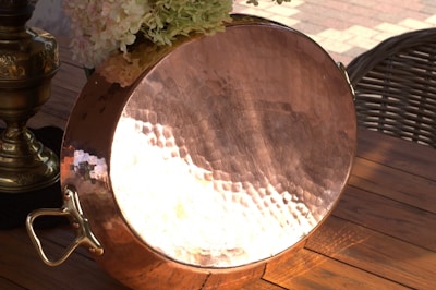 A hammered copper pot with a shiny, reflective surface is placed on a wooden table. In the background, there is a brass decorative item and a bouquet of hydrangeas.