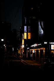 A dimly lit street scene at night with illuminated signs in Korean. Several storefronts are visible, with warm lights glowing from one establishment. A few people are walking along the sidewalk, and shadows dominate the scene.