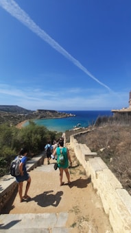 A group of people are walking down a stone path towards a beautiful coastal area. The sea is a vivid blue-green, and the sky is mostly clear with a single white contrail stretching across it. Sparse foliage lines the path, and the terrain appears rugged and natural.