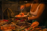A person serves a plate of food from a selection of trays containing various cooked dishes. The setting appears to be a buffet with trays of food including shrimp and other items. Tongs are being used to pick the food, and the area is dimly lit.