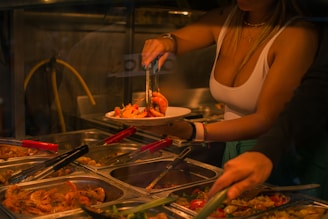 A person serves a plate of food from a selection of trays containing various cooked dishes. The setting appears to be a buffet with trays of food including shrimp and other items. Tongs are being used to pick the food, and the area is dimly lit.
