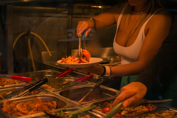 A person serves a plate of food from a selection of trays containing various cooked dishes. The setting appears to be a buffet with trays of food including shrimp and other items. Tongs are being used to pick the food, and the area is dimly lit.