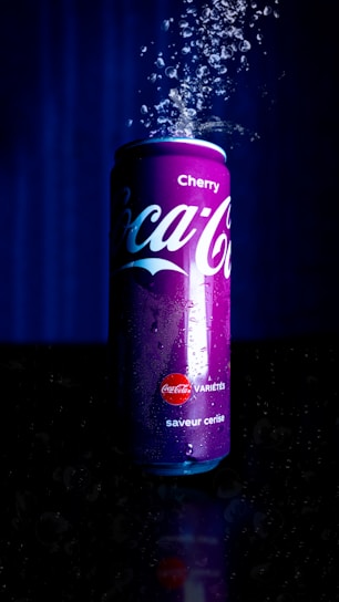 A can of Cherry Coca-Cola is surrounded by rising bubbles, illuminated against a dark background. The can is red and prominently features the brand logo and the flavor indication. Water droplets are visible on its surface, enhancing the refreshing appeal.