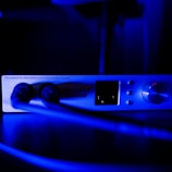 A close-up view of an audio interface device labeled 'Discrete 4 Synergy Core.' The device is connected with two cables and illuminated in blue light, emphasizing the knobs, buttons, and screen on its front panel.
