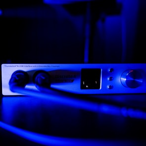 A close-up view of an audio interface device labeled 'Discrete 4 Synergy Core.' The device is connected with two cables and illuminated in blue light, emphasizing the knobs, buttons, and screen on its front panel.
